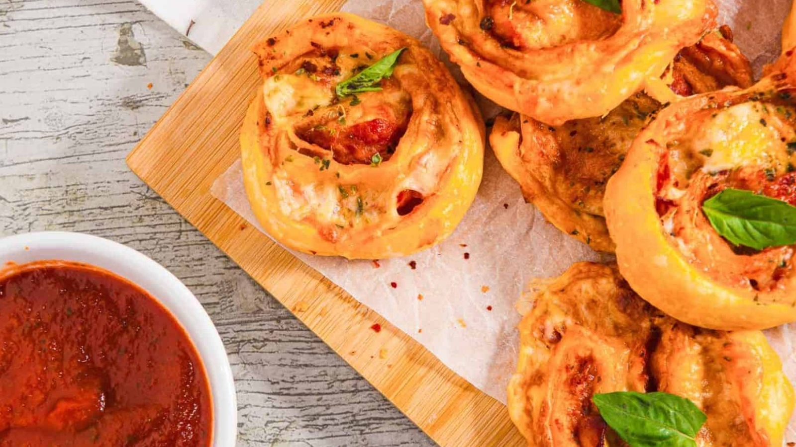 A wooden board holds several pizza rolls topped with basil leaves, set on parchment paper. A small bowl of marinara sauce is on the left. The table surface is light gray with visible grain patterns.