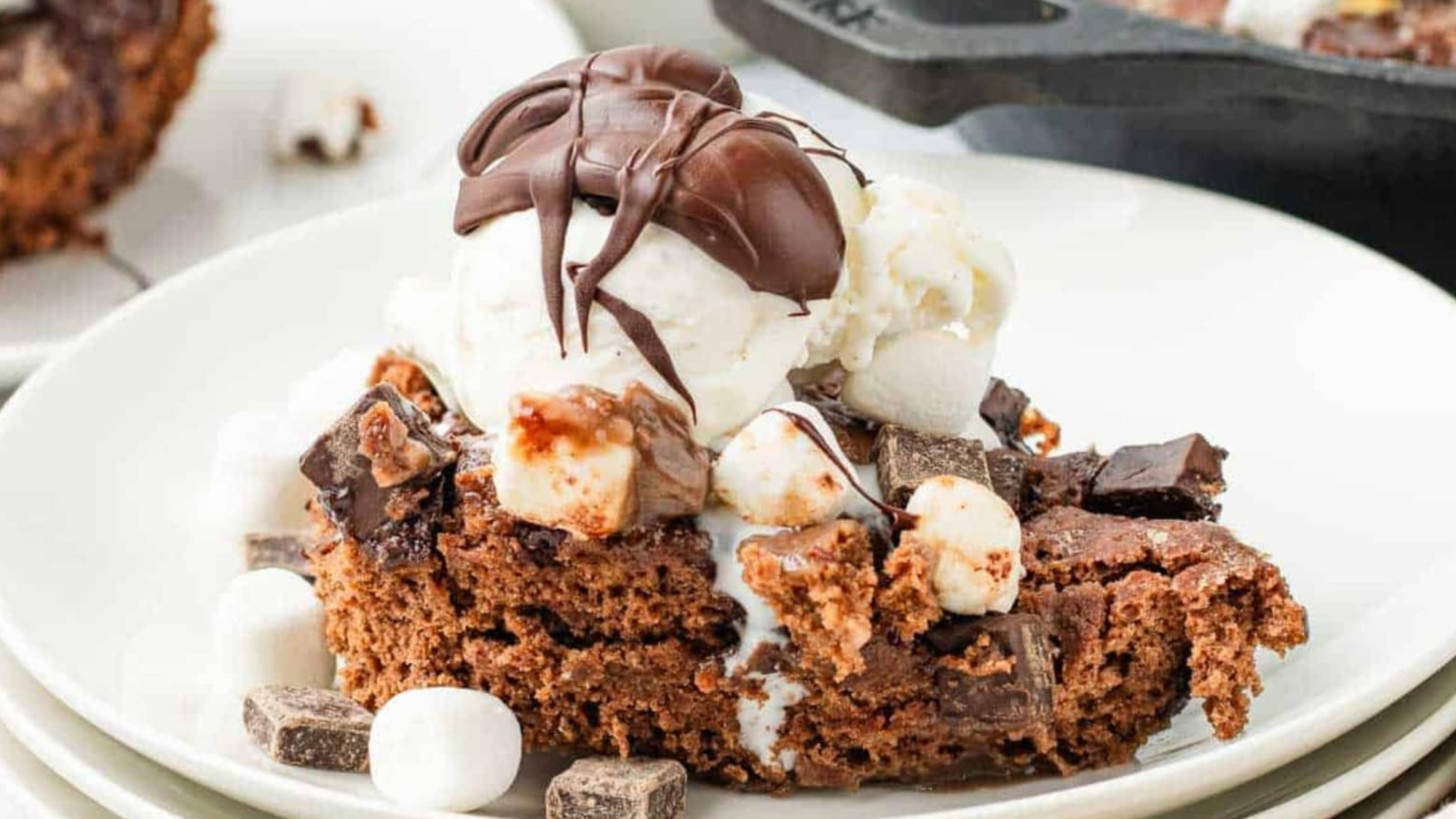 A chocolate brownie topped with ice cream, marshmallows, and chocolate drizzle on a white plate.