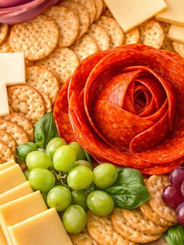 Charcuterie platter with meat roses made from pepperoni arranged with crackers, cheese, grapes, and basil.