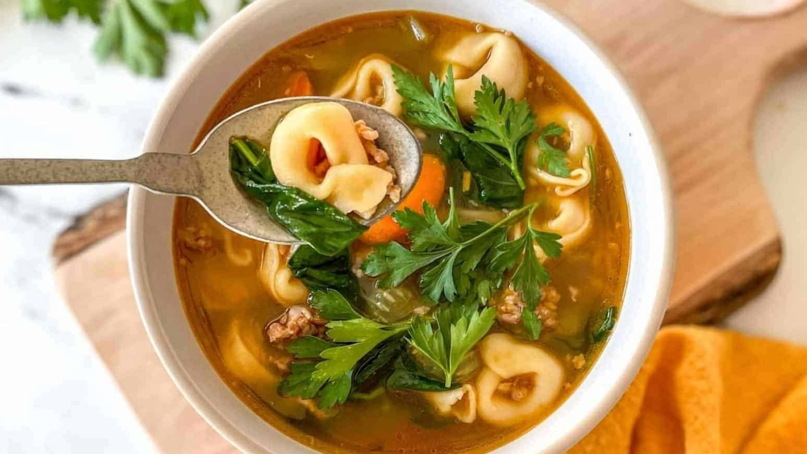 A bowl of tortellini soup sits on a rustic wooden cutting board with a yellow linen and a bunch of parsley.
