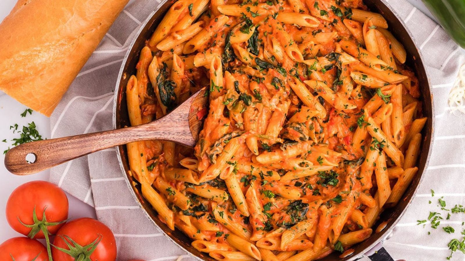 Penne pasta in a creamy pink sauce fills a skillet with a wooden spoon resting inside, surrounded by tomatoes, bread, cheese, and herbs on a cloth surface.