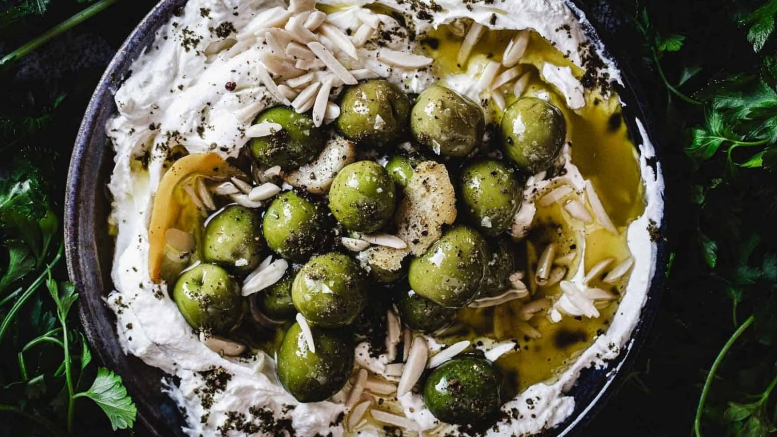 A bowl of whipped labneh topped with green olives, olive oil, slivered almonds, and herbs, surrounded by fresh parsley leaves.