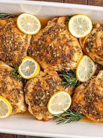 Baked chicken thighs in a white dish, seasoned with herbs and lemon slices, garnished with rosemary sprigs.
