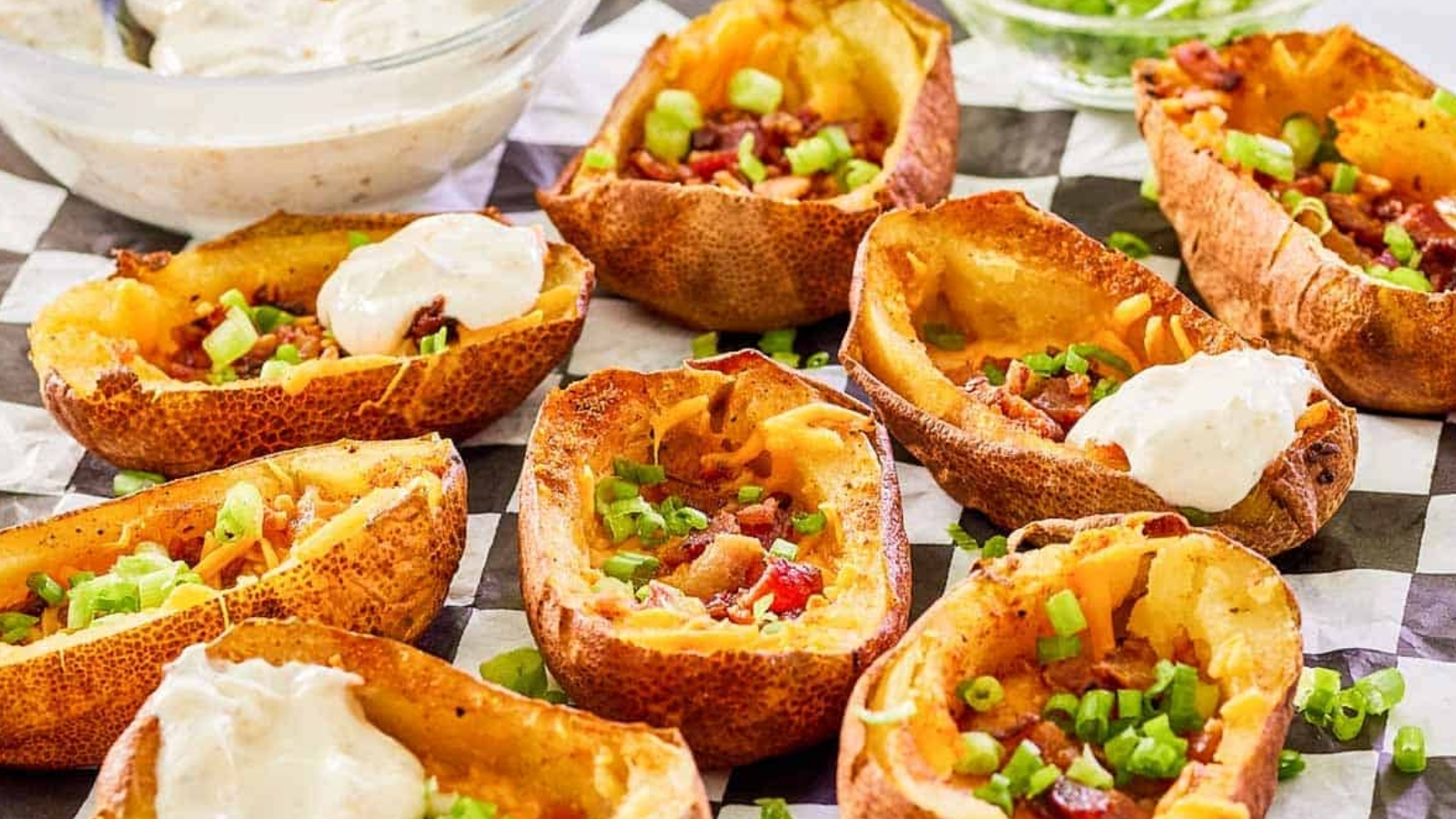 Halved baked potatoes filled with cheese, bacon pieces, and chopped green onions are arranged on checkered paper with bowls of sour cream and sliced green onions in the background.