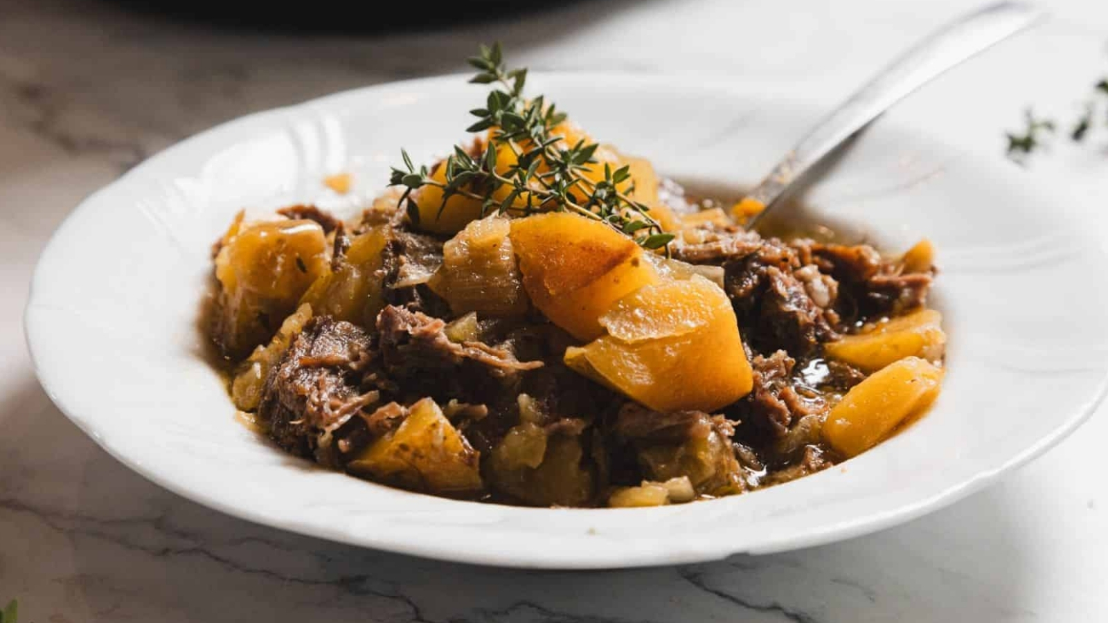A white plate containing a stew made of tender shredded meat and chunks of potatoes, garnished with sprigs of fresh thyme. A spoon rests on the side of the dish, placed on a marble surface.