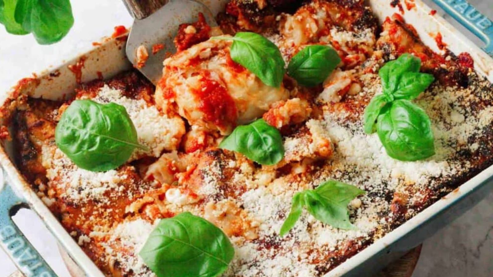 A baked lasagna in a rectangular dish, topped with tomato sauce, basil leaves, and grated cheese. A spatula is lifting a portion from the dish, revealing layers of pasta and filling. The dish is on a light countertop.