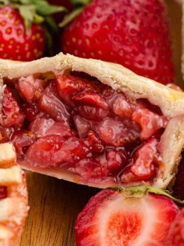 A bitten mini strawberry pie with visible filling sits on a wooden surface beside whole and halved strawberries.