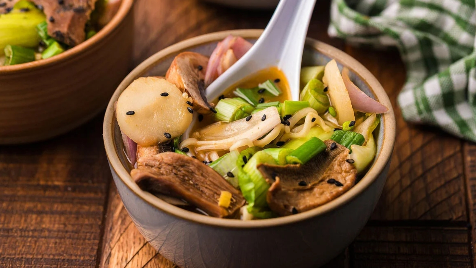 A bowl of noodle soup with sliced mushrooms, green onions, and black sesame seeds sits on a wooden surface with a white spoon resting inside.