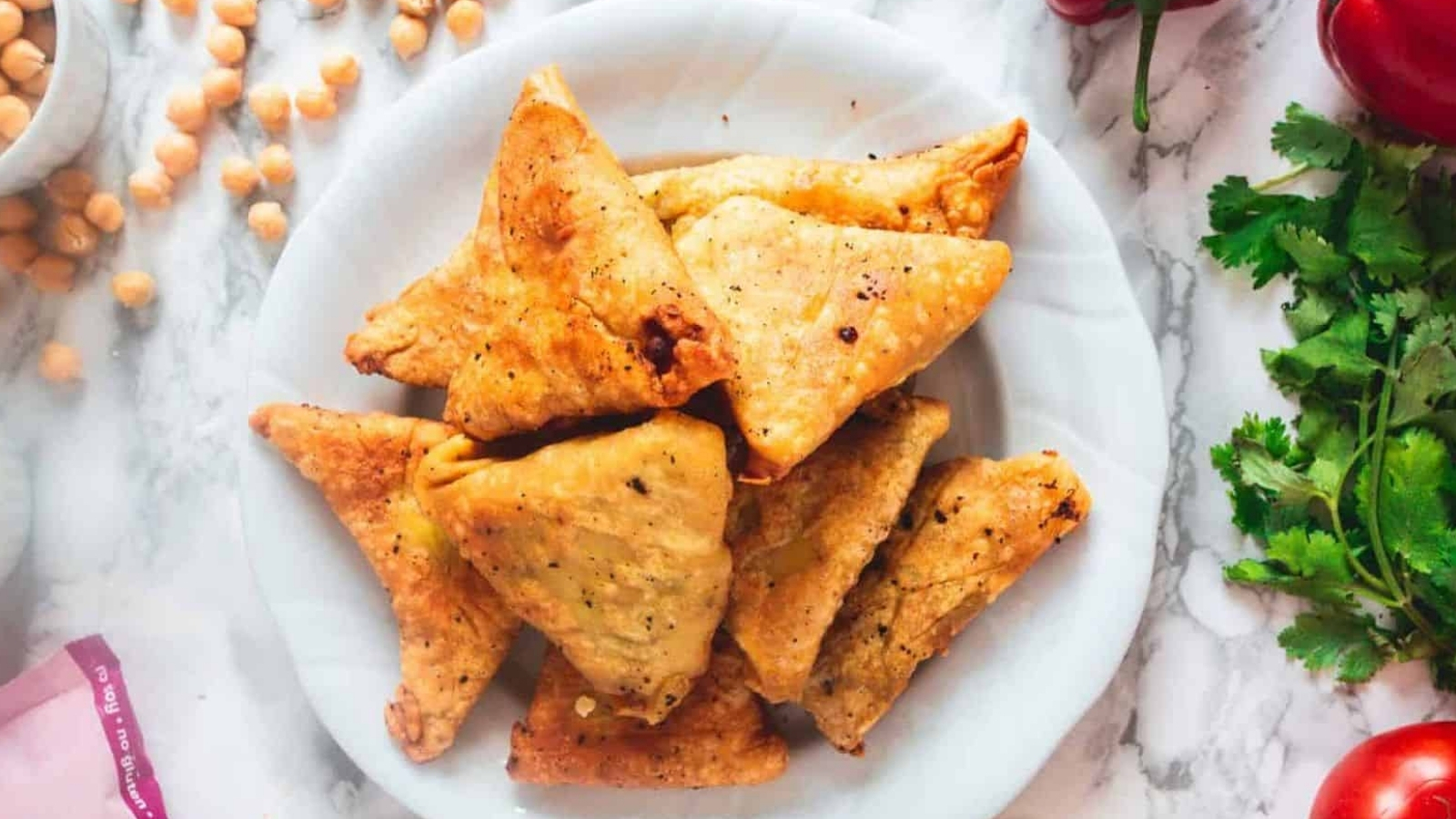 A plate of golden-brown triangular samosas is placed on a marble surface. Surrounding the plate are scattered chickpeas, fresh cilantro, a red pepper, and a cutting board corner. The samosas appear crispy and seasoned.