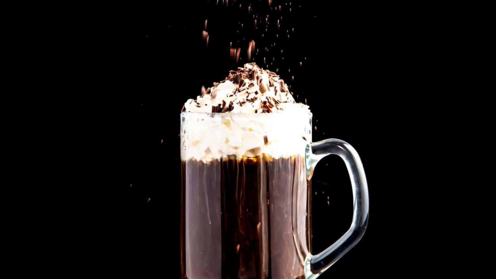 Glass mug of hot chocolate topped with whipped cream and chocolate shavings against a black background.