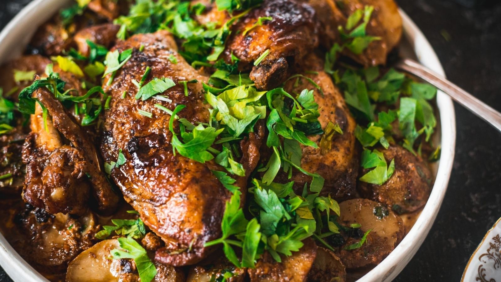 A platter of roasted chicken garnished with fresh parsley, accompanied by sautéed mushrooms and potato slices, with a spoon next to the dish. A bowl of creamy white sauce is visible in the background.