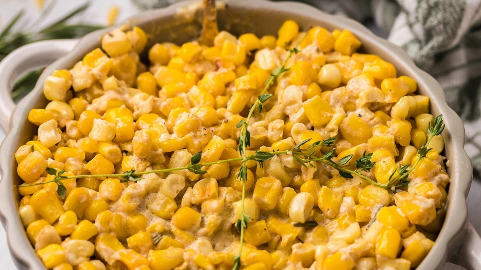 Round dish filled with creamed corn and diced yellow pieces, topped with sprigs of herbs.