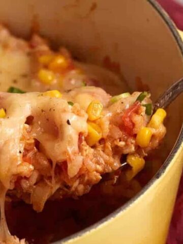 A spoon scoops cheesy rice, corn, and vegetables from a yellow pot with melted cheese on top.