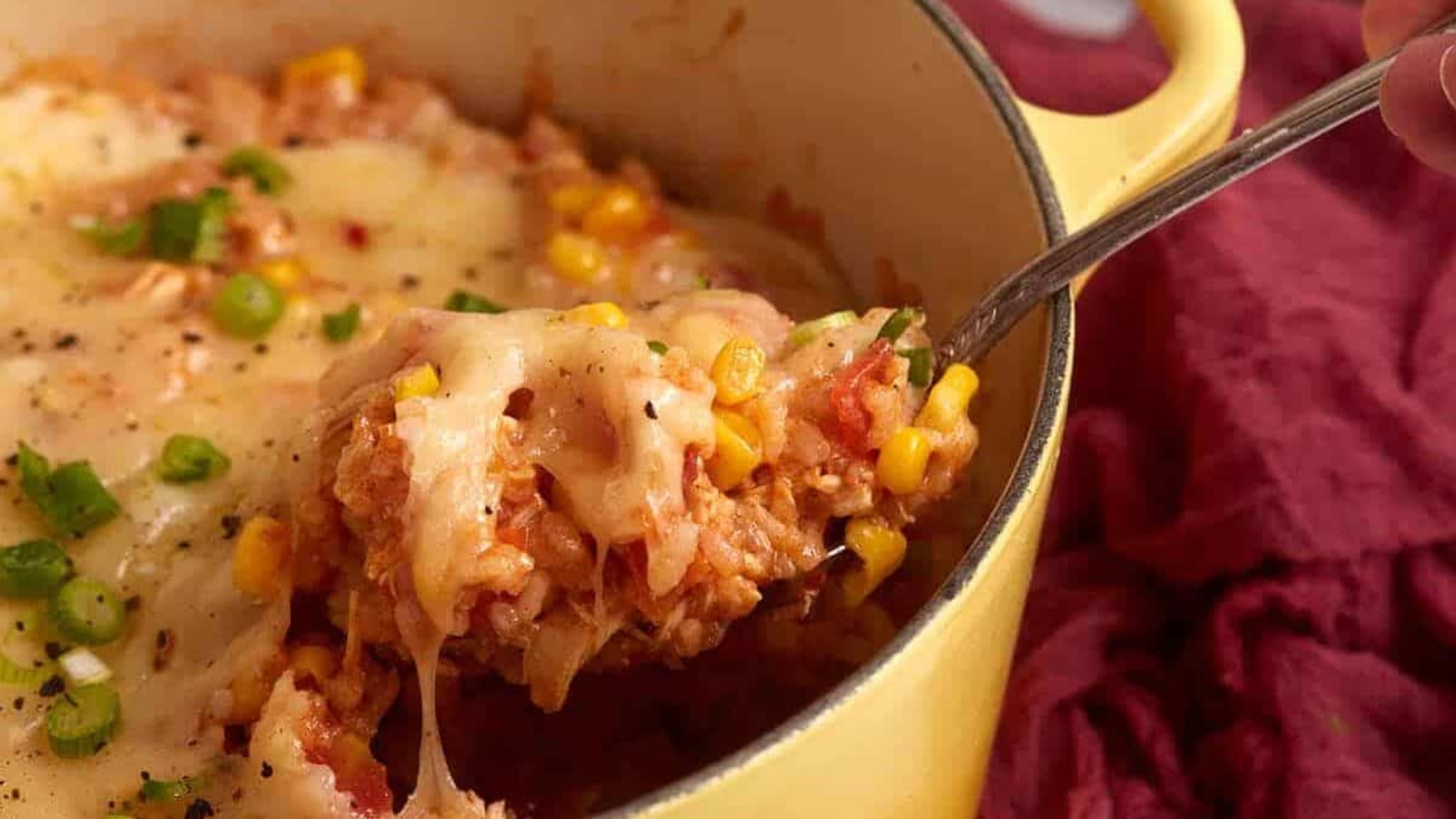 A spoon scoops cheesy rice, corn, and vegetables from a yellow pot with melted cheese on top.