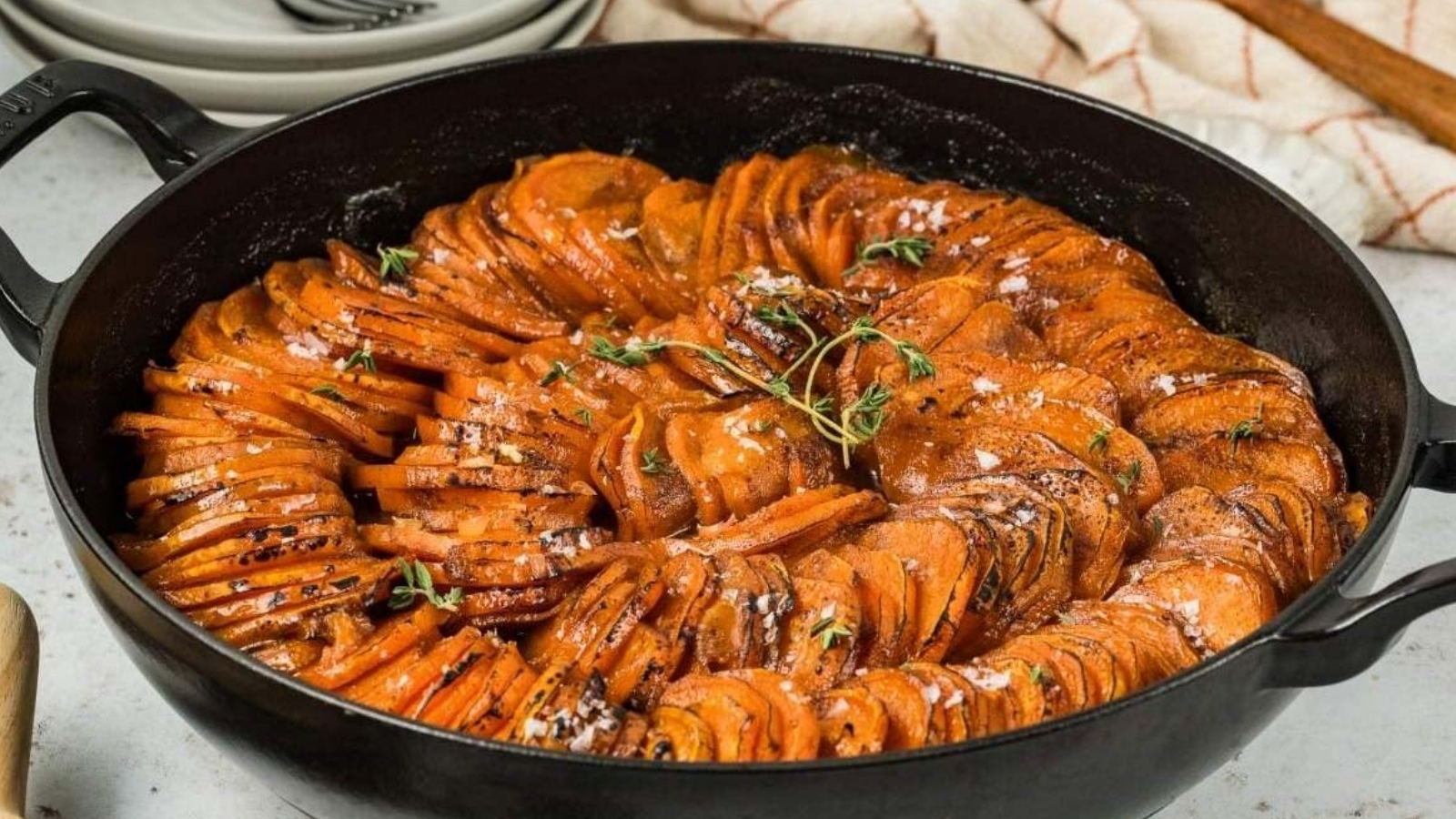 Thinly sliced sweet potatoes are arranged in a circular pattern in a black skillet and topped with herbs and coarse salt.
