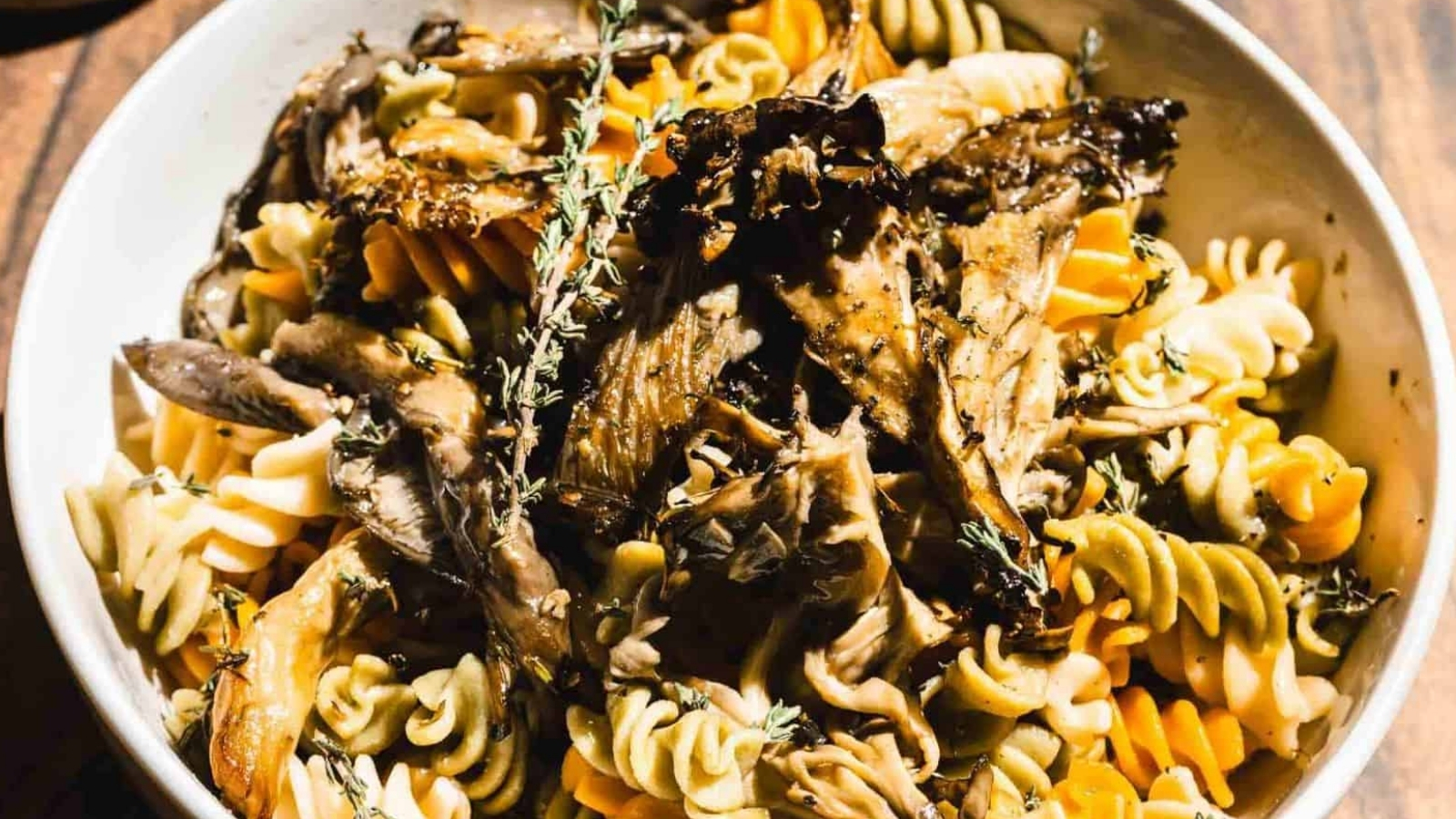 A bowl of multicolored fusilli pasta topped with cooked mushrooms and garnished with fresh thyme sprigs. The dish is served in a white bowl on a wooden surface.