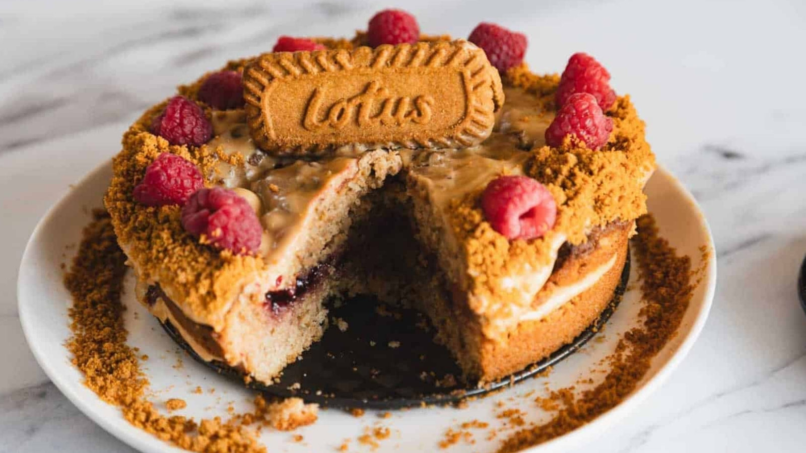 A decorated cake with raspberries and Lotus biscuit, with a slice removed, on a white plate.