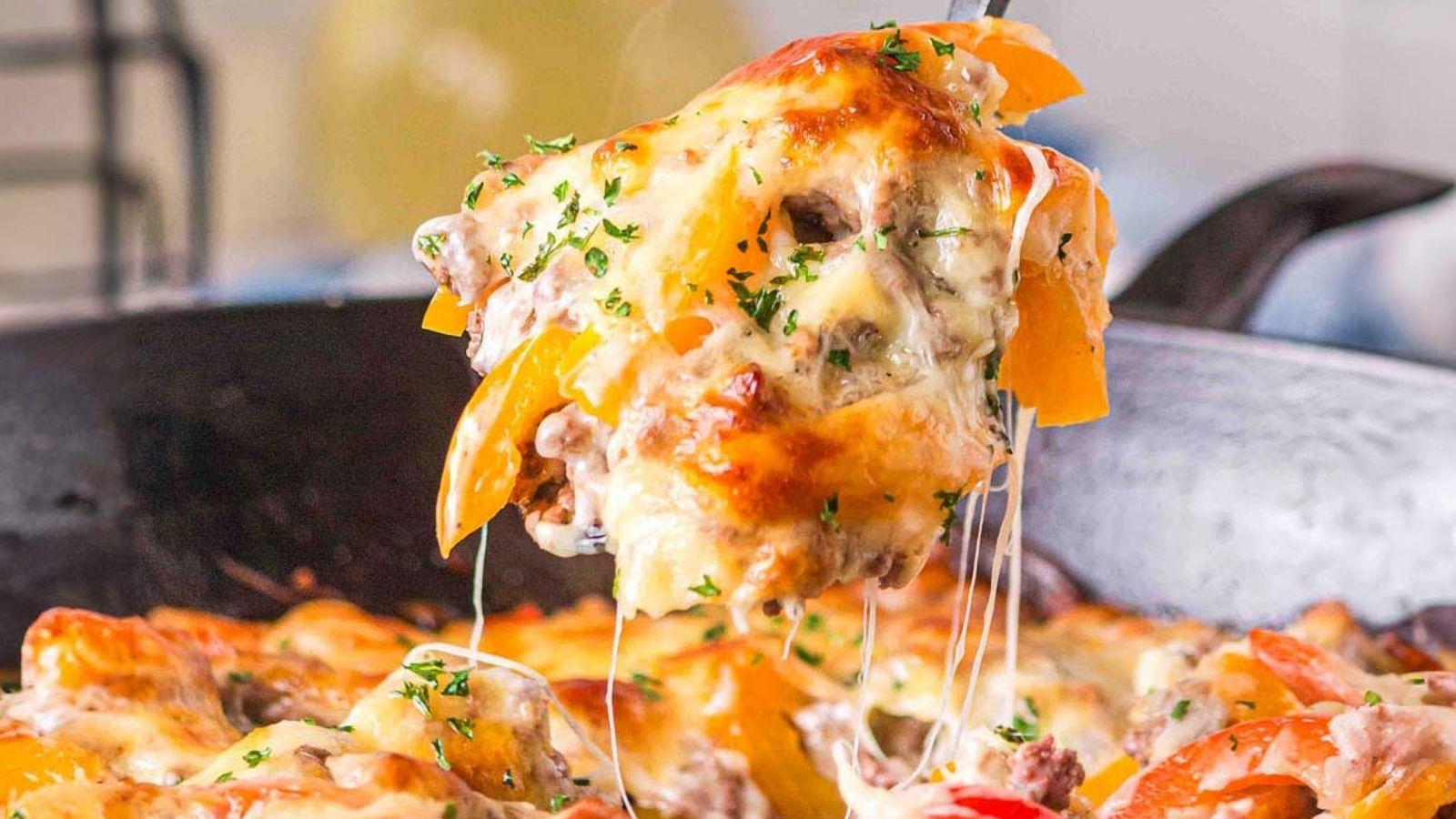 A spoon lifts a serving of baked pasta with melted cheese, ground meat, mushrooms, and sliced bell peppers from a skillet.
