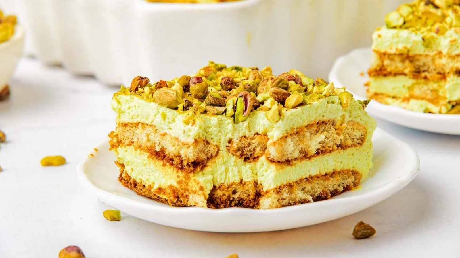 A slice of pistachio tiramisu topped with chopped pistachios on a white plate. Layers of cream and sponge cake are visible.