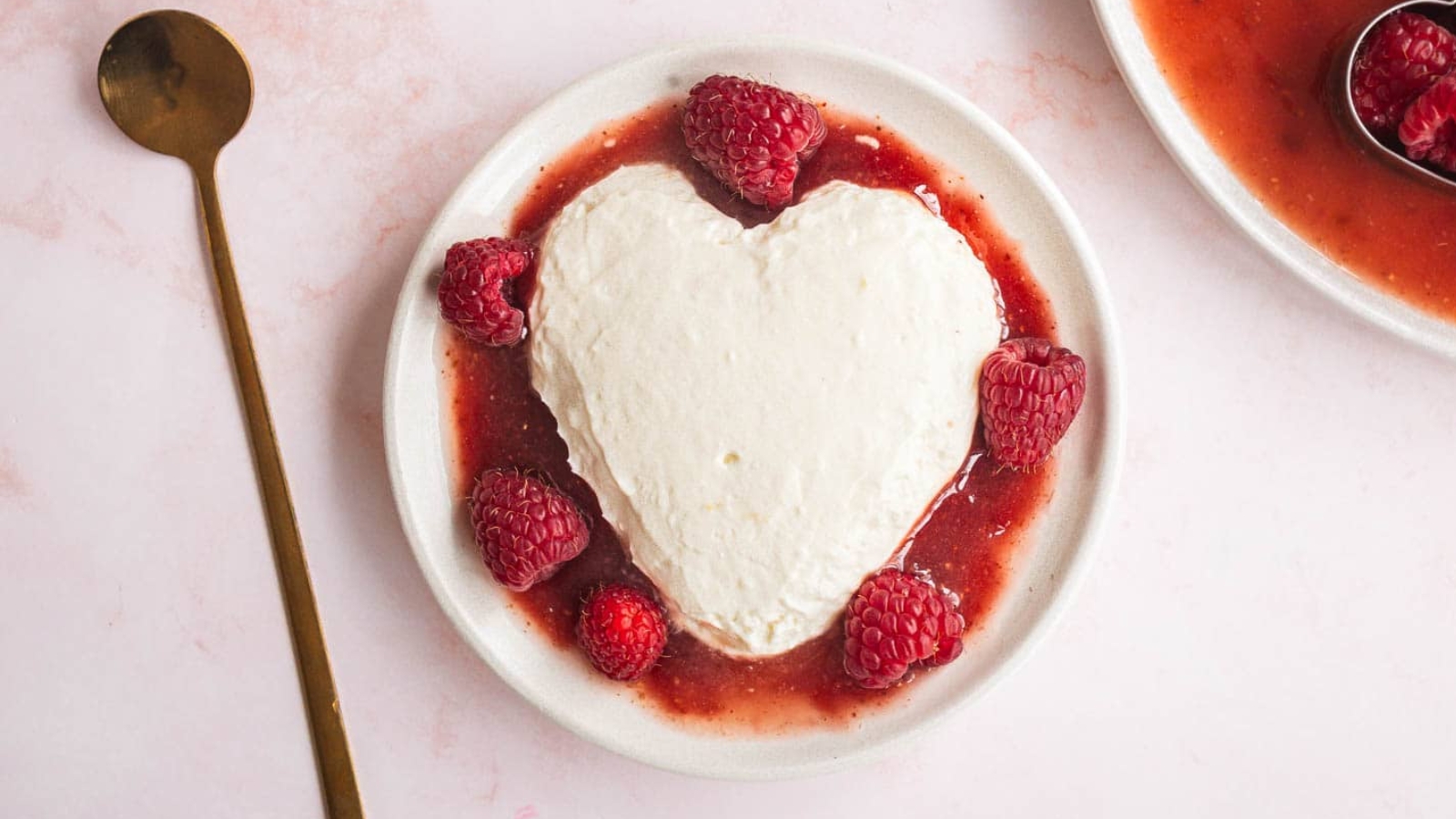 A heart-shaped dessert covered in a white cream sits on a plate with a raspberry sauce and whole raspberries. A person holds a small gold spoon just above the dessert. Another heart-shaped dessert with similar presentation is partially visible in the background.
