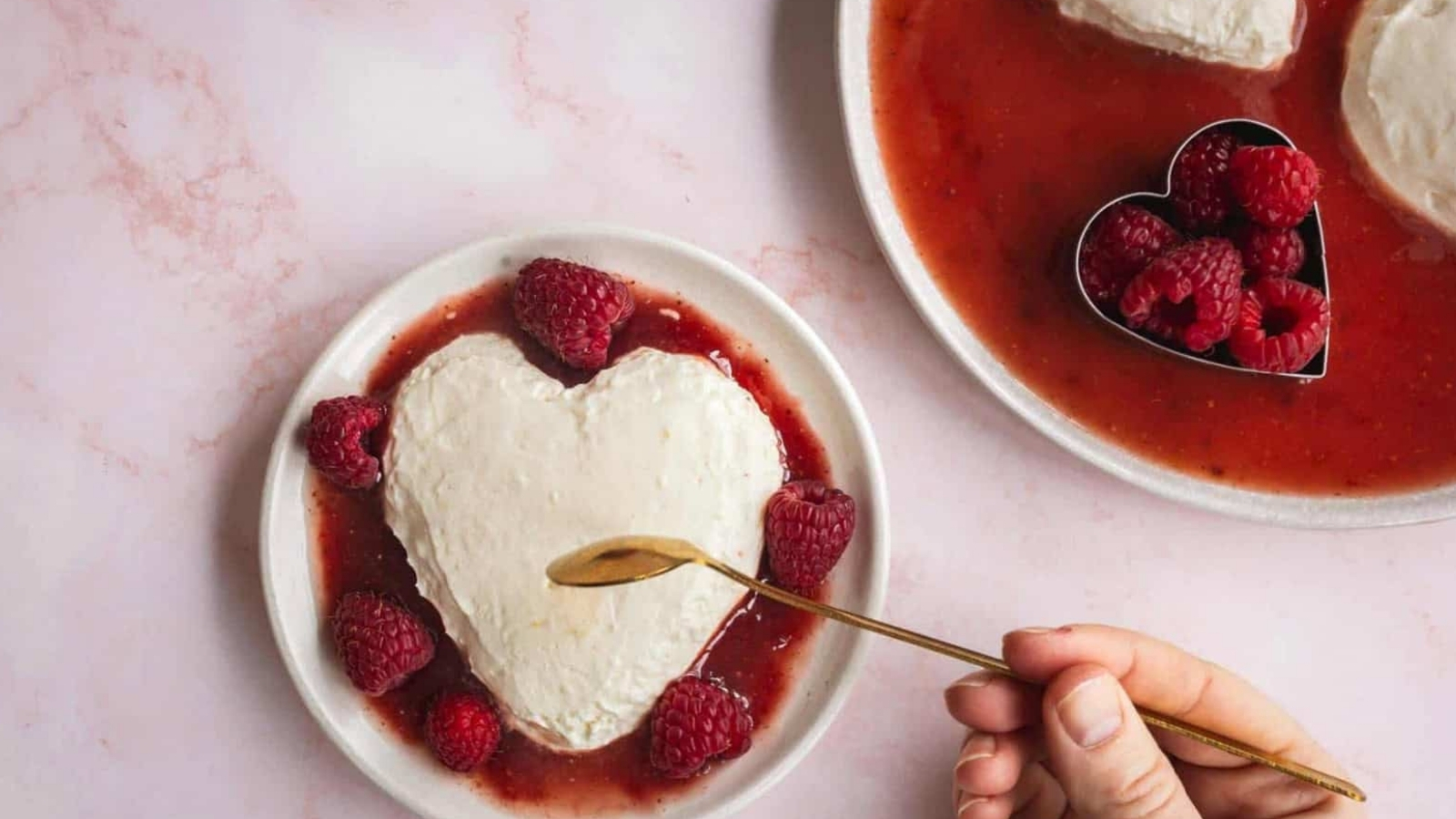 A heart-shaped dessert covered in a white cream sits on a plate with a raspberry sauce and whole raspberries. A person holds a small gold spoon just above the dessert. Another heart-shaped dessert with similar presentation is partially visible in the background.