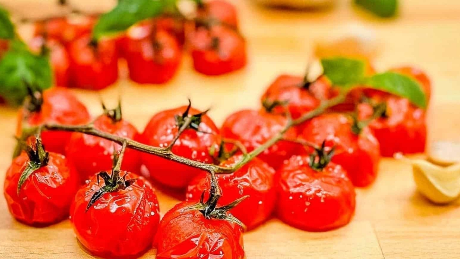 A closeup shot of roasted cherry tomatoes on the vine on a wooden cutting board surrounded by basil and garlic.