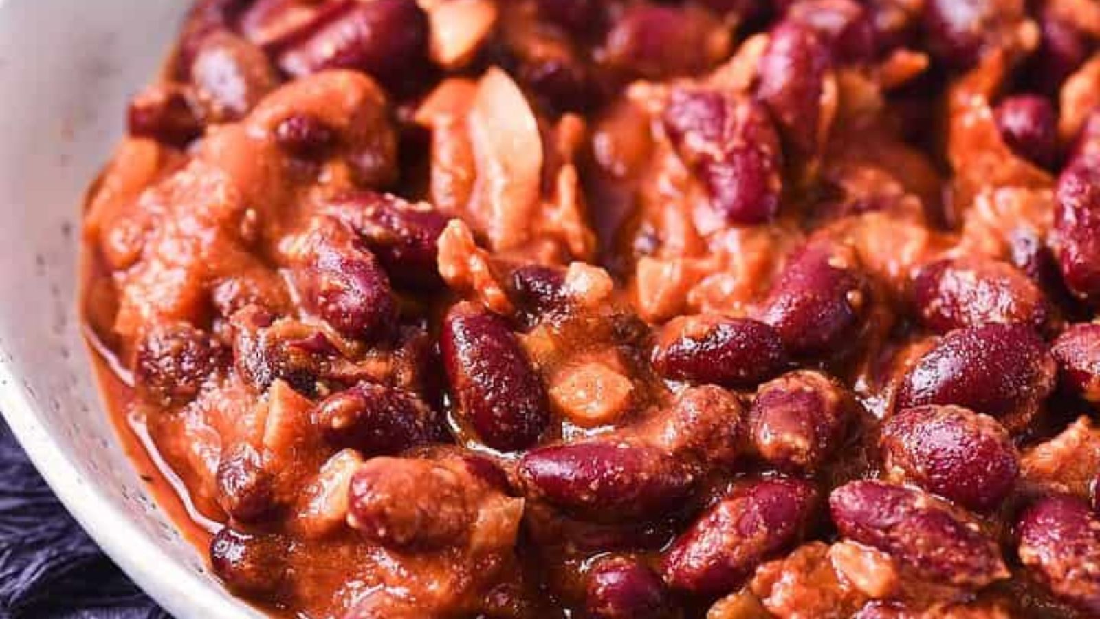 A white bowl filled with a red kidney bean stew, including visible pieces of meat or vegetables, sits on a dark cloth napkin with a fork and spoon beside it. A small bunch of parsley is on the table nearby.