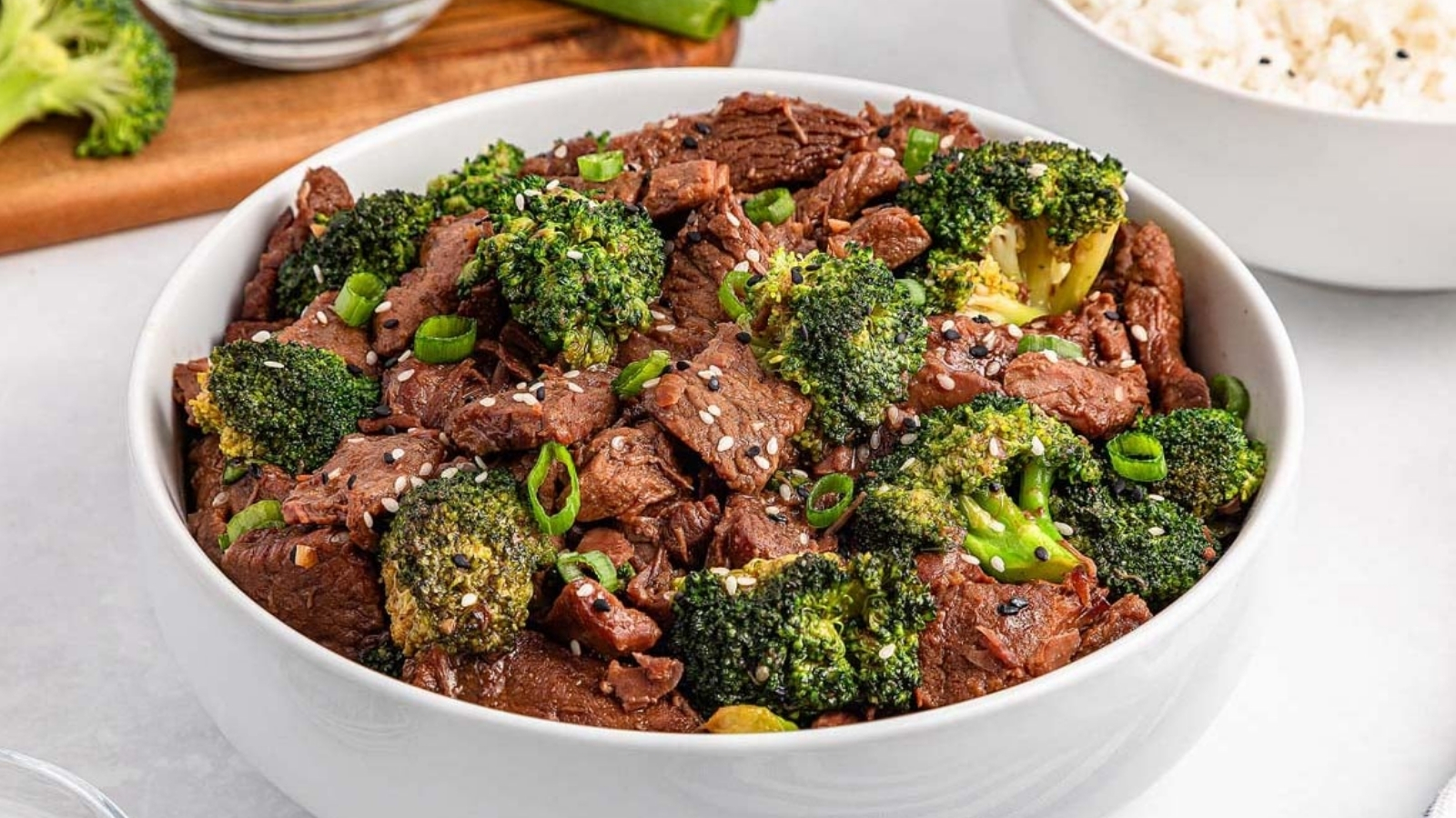 A white bowl filled with sliced beef and broccoli florets topped with sesame seeds and chopped green onions sits on a light surface, with a bowl of rice in the background.