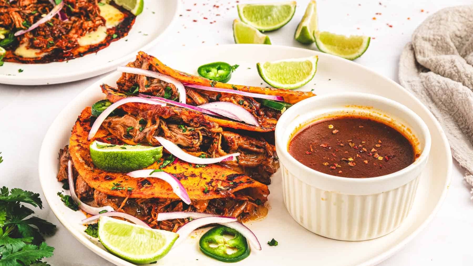 Three birria tacos with lime wedges, sliced onions, and dipping sauce on a white plate.