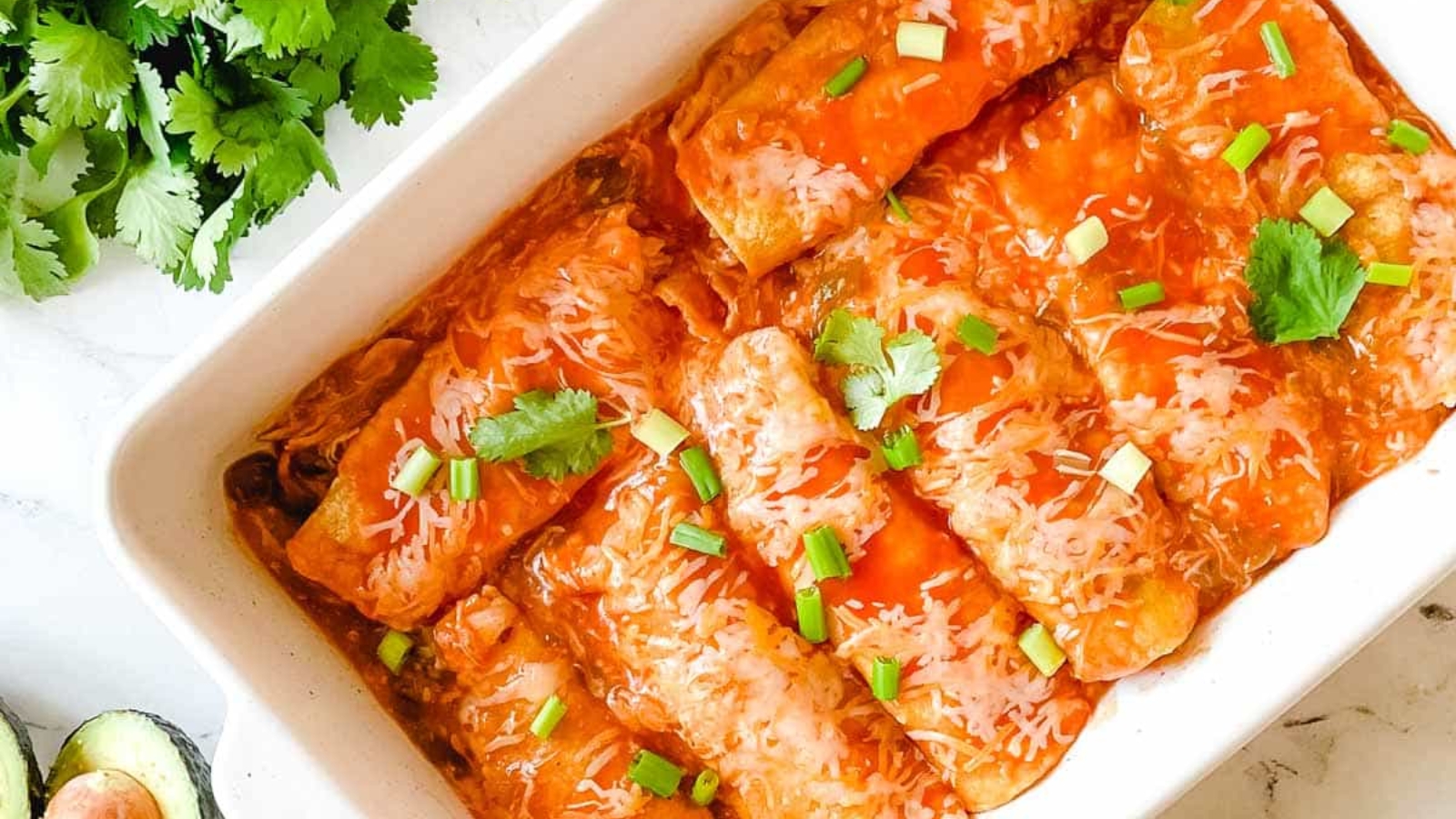 A baking dish full of chicken enchiladas is surrounded by cut limes, a halved avocado, and a green linen.