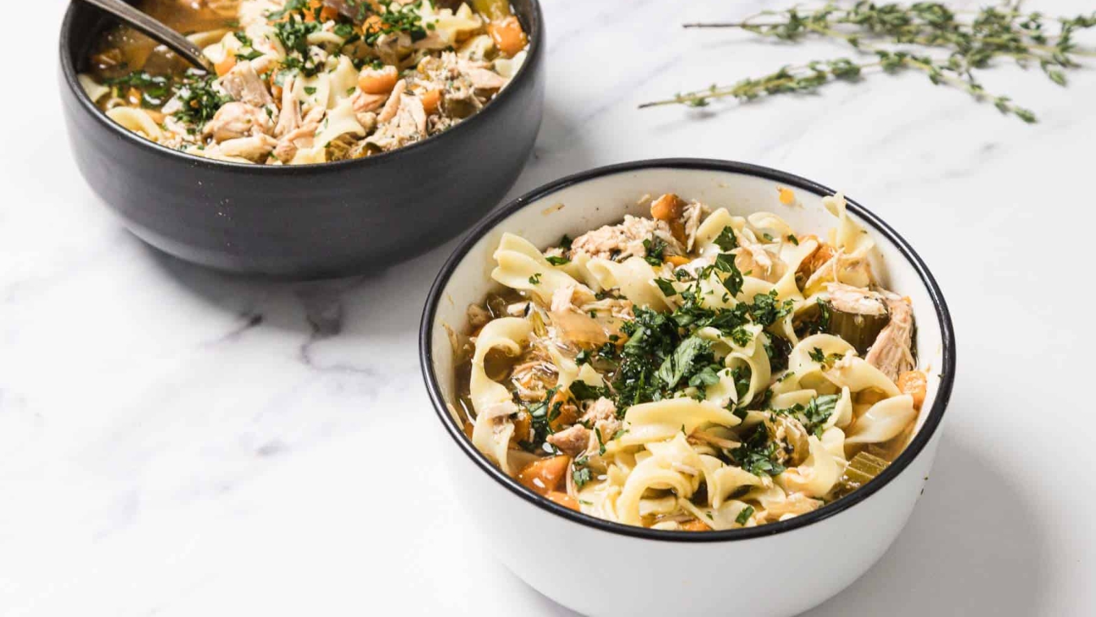 A bowl of chicken noodle soup with carrots, celery, herbs, and a spoon inside.