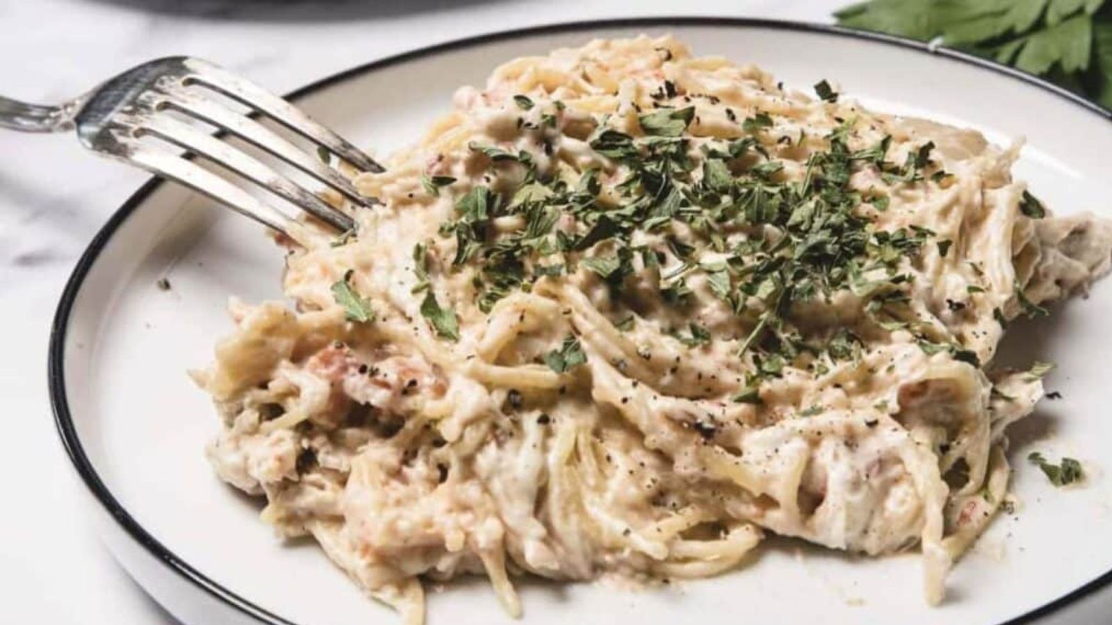 A plate of creamy spaghetti with shredded chicken, topped with chopped parsley and black pepper.