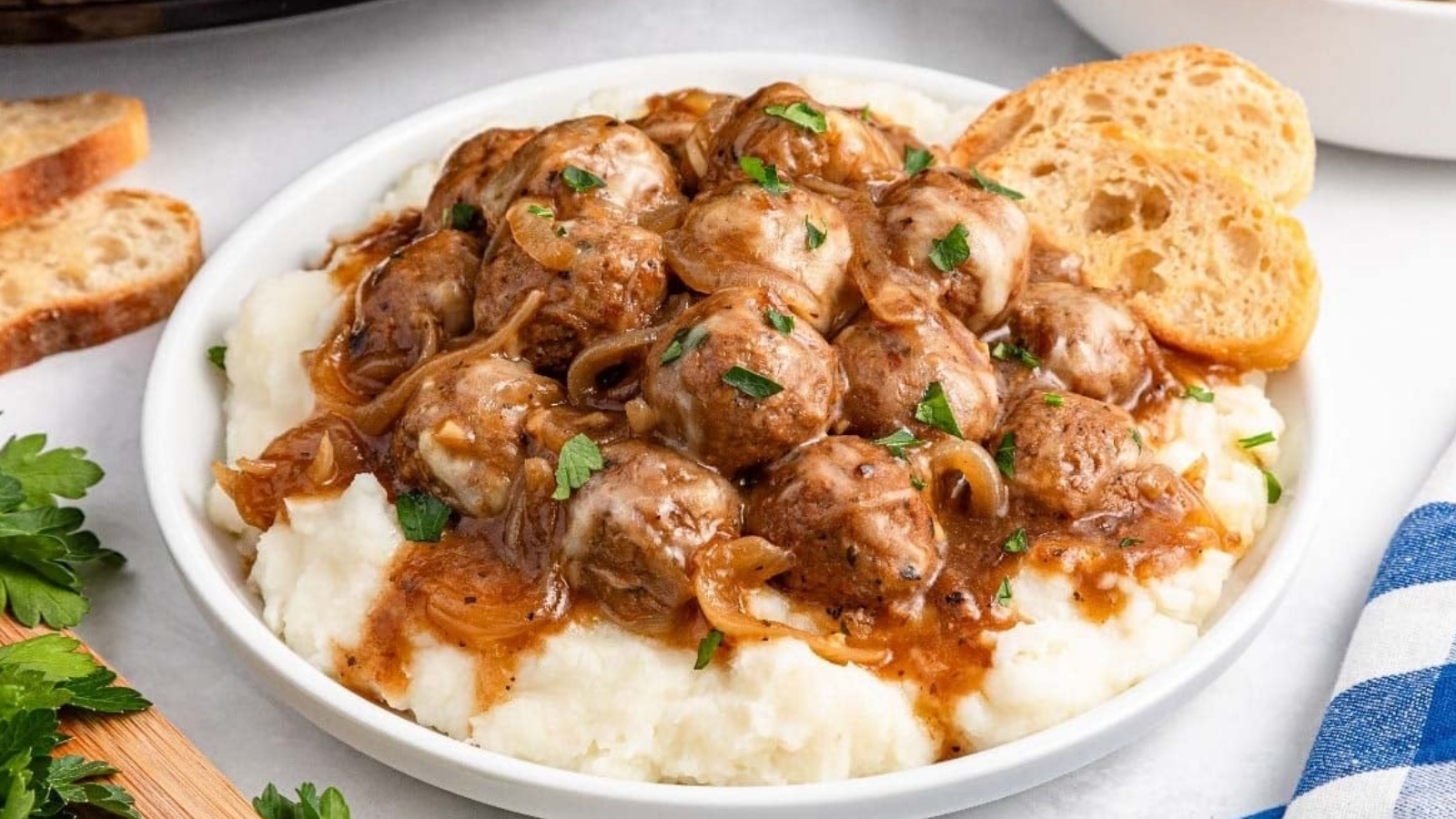 Meatballs with brown gravy and sliced onions are served over mashed potatoes in a bowl, topped with chopped herbs and accompanied by slices of bread.