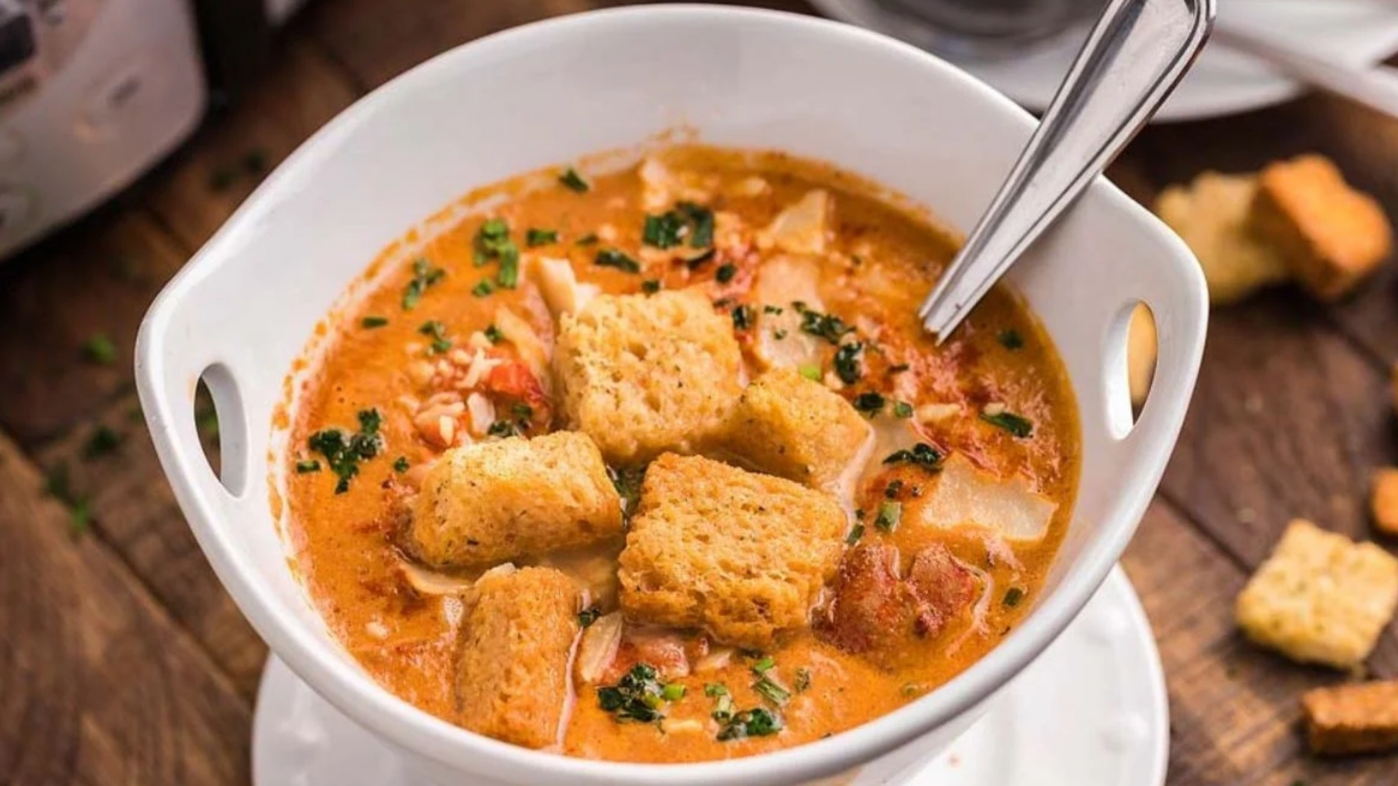 A white bowl of creamy tomato-based soup topped with croutons and chopped herbs, with a spoon inside, set on a wooden surface with extra croutons nearby.