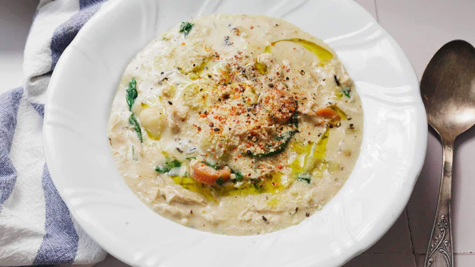 Creamy soup with vegetables and herbs in a white bowl, next to a spoon and a striped towel.