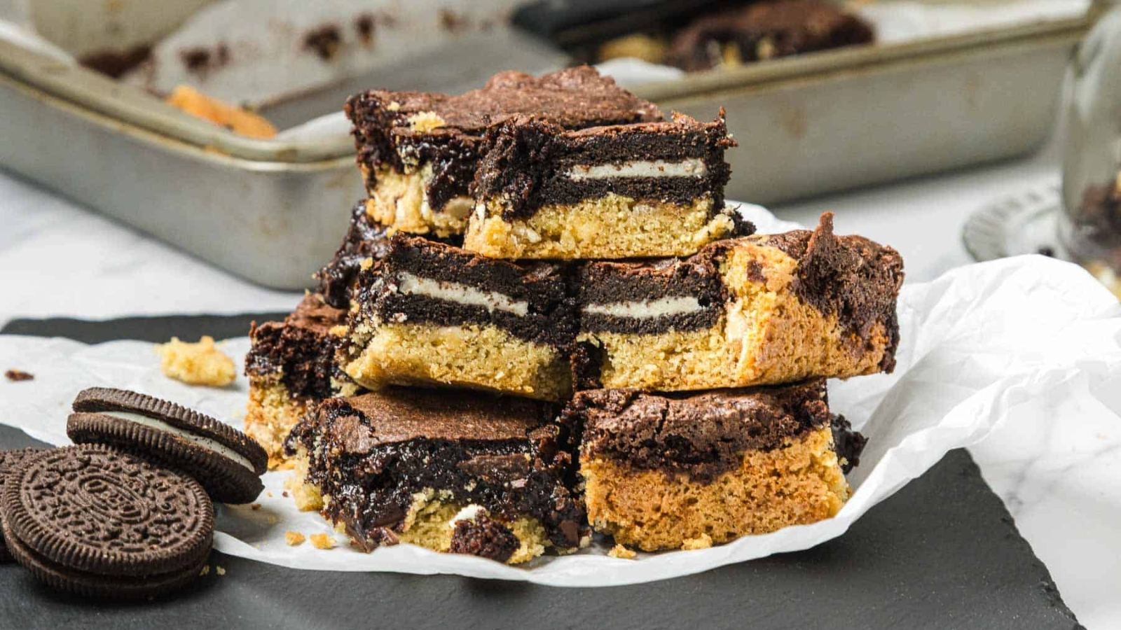 A stack of layered cookie and brownie bars with Oreo cookies inside, displayed on parchment paper.