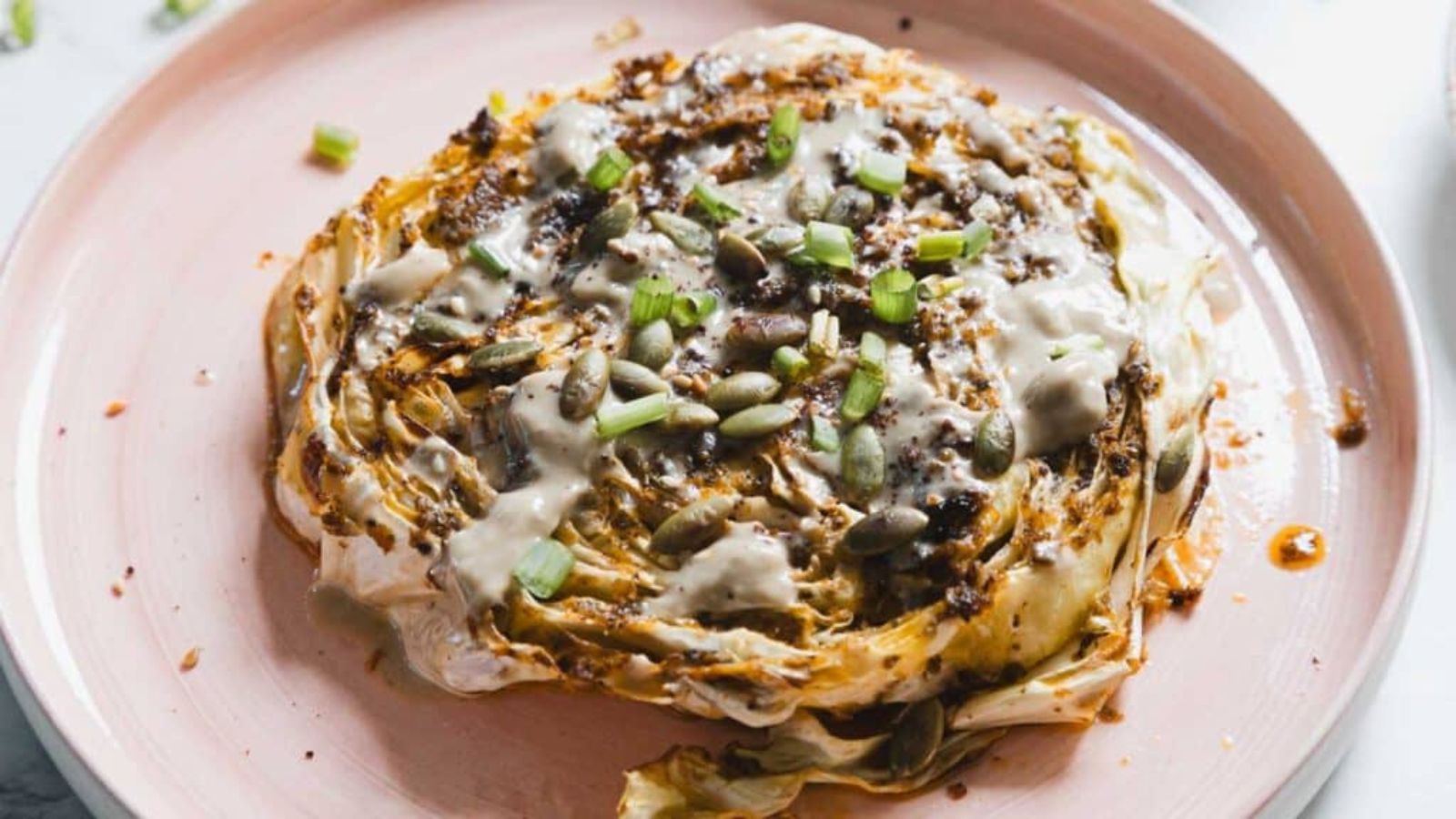 A roasted cabbage steak is served on a pink plate. It is topped with a creamy sauce, pumpkin seeds, and chopped green onions. The cabbage is browned and slightly charred at the edges.