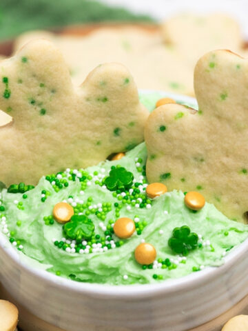 Shamrock-shaped cookies around a small bowl of green frosting topped with green and white sprinkles and gold candy pieces.