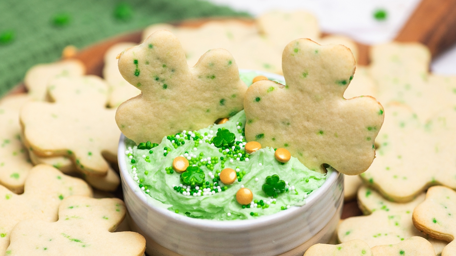 Shamrock-shaped cookies around a small bowl of green frosting topped with green and white sprinkles and gold candy pieces.