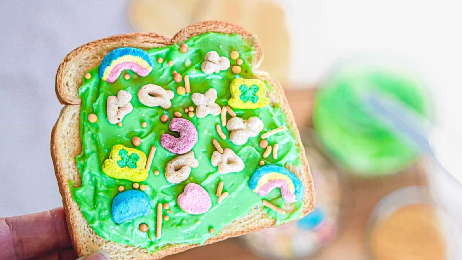 A slice of toast spread with bright green frosting is topped with colorful marshmallow cereal pieces and small sprinkles. A jar of green frosting, a bowl of cereal, and a knife are blurred in the background.