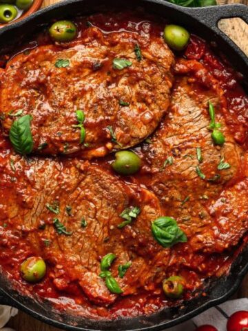 Steak slices in a cast-iron pan covered with chunky tomato sauce and green olives, surrounded by small bowls of herbs and sliced bread.