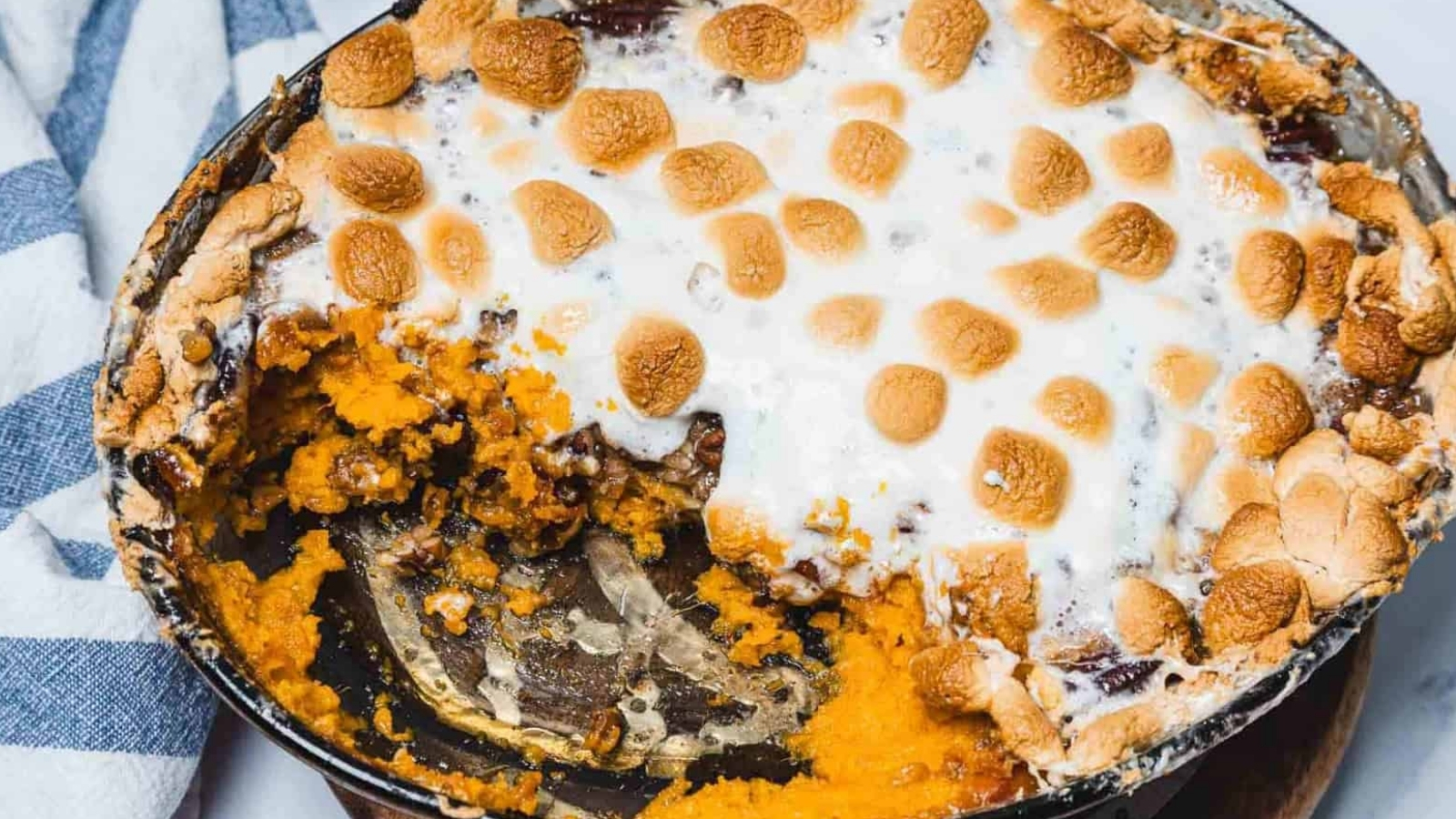 Baked sweet potato casserole topped with toasted marshmallows in a round dish, partially eaten, with a metal serving spoon visible.