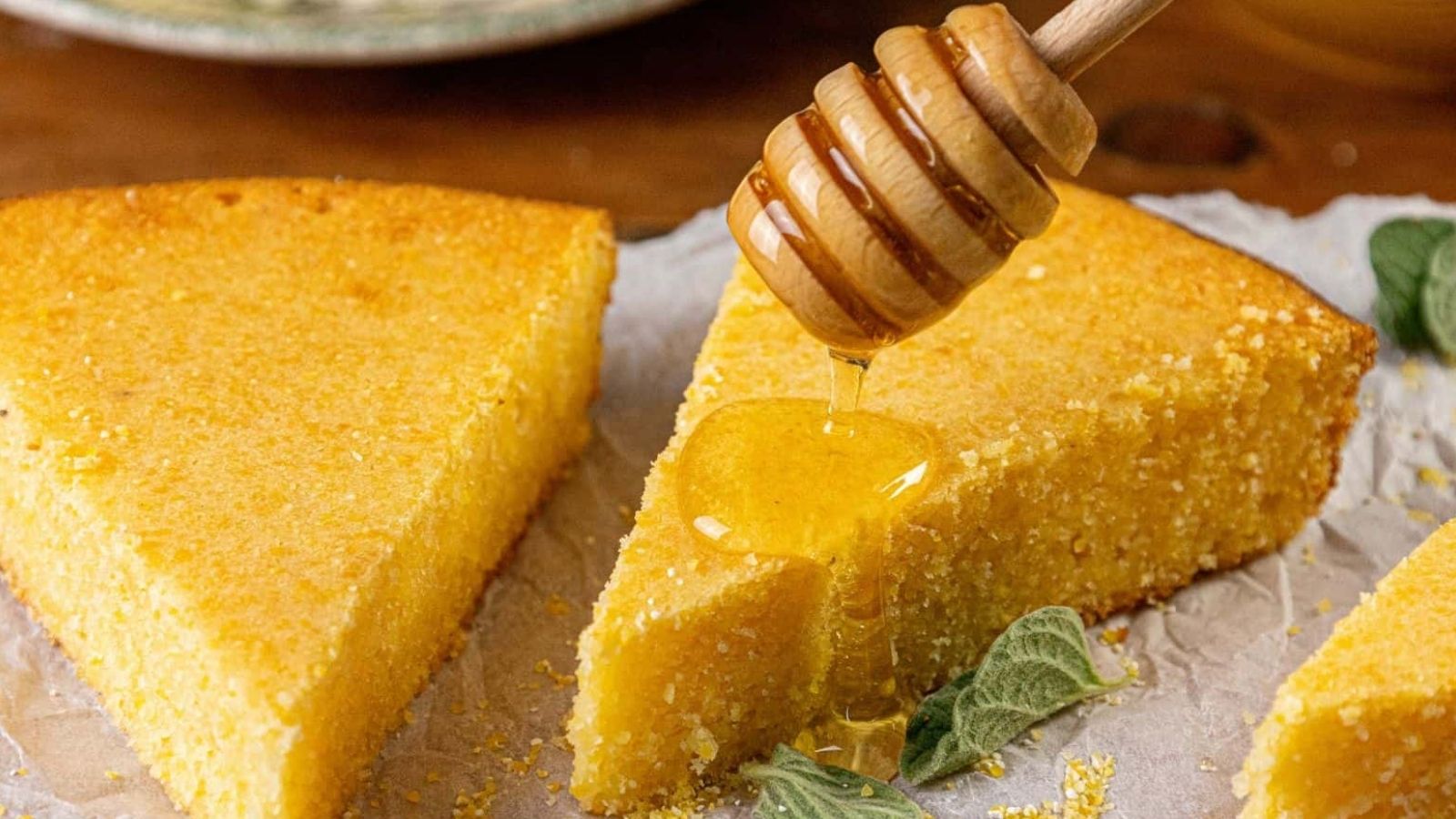 Close-up of honey being drizzled onto a slice of Southern cornbread with a wooden honey dipper. The cornbread, boasting a moist center and golden crust, sits on parchment paper, accompanied by green leaves. The softly blurred background highlights its inviting texture.