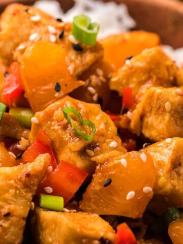 Chunks of chicken with orange segments and red bell pepper pieces topped with sliced green onions and sesame seeds over white rice in a bowl, with orange wedges on the side.