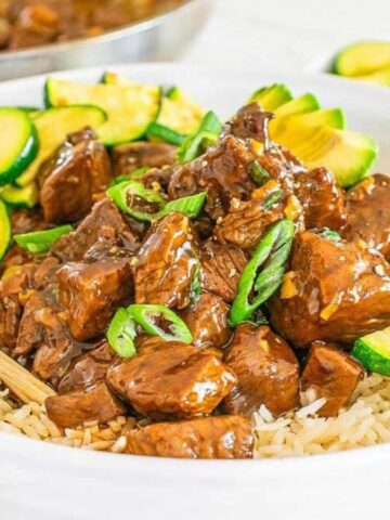 A bowl of rice topped with cubed steak bites in a brown sauce, garnished with sliced green onions and zucchini. .