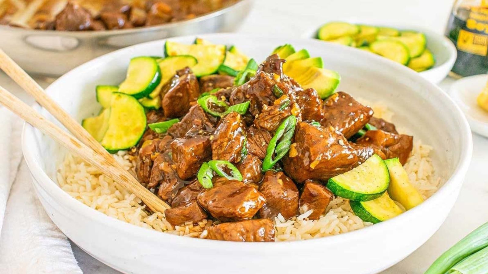 A bowl of rice topped with cubed steak bites in a brown sauce, garnished with sliced green onions and zucchini.