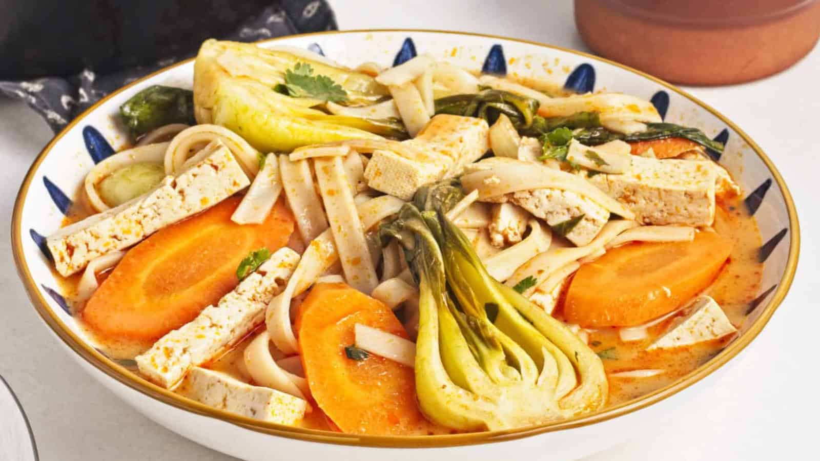 A bowl of noodle soup with tofu slices, baby bok choy, carrot rounds, and fresh herbs in a light, orange-colored broth.