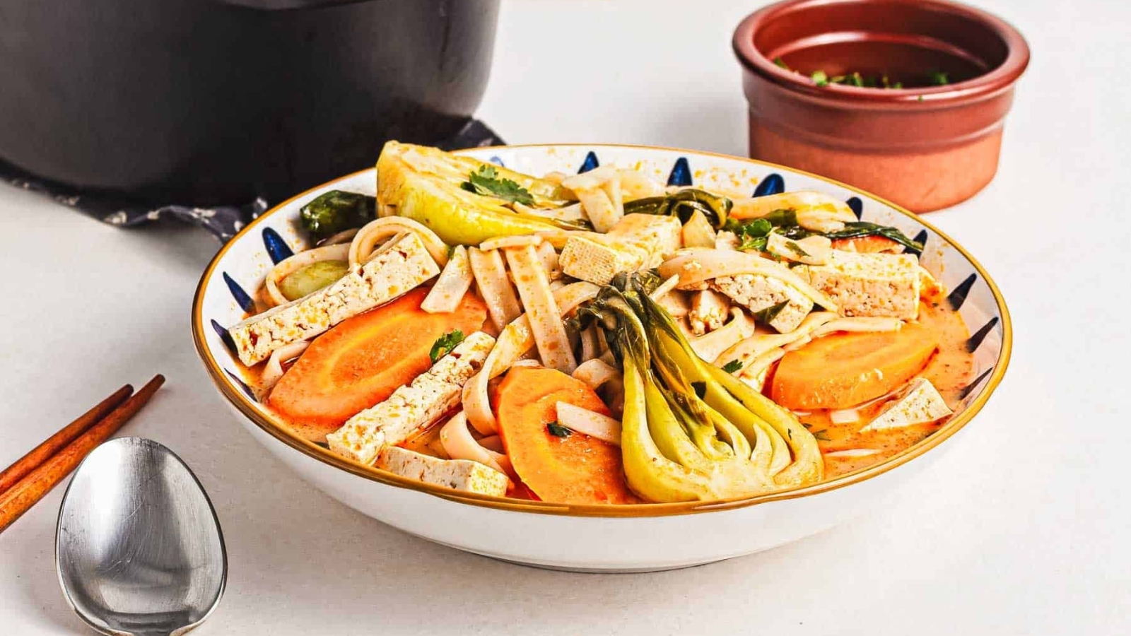 A bowl of noodle soup with tofu strips, baby bok choy, carrot slices, and fresh herbs, set on a white surface with a spoon nearby and a small bowl of chopped greens in the background.