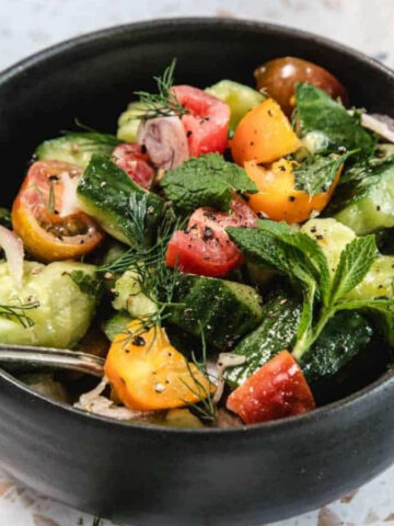 A black bowl filled with a salad made of sliced cucumbers, cherry tomatoes in various colors, fresh herbs, and a fork resting on the side. A white cloth with blue stripes is partially visible in the background.