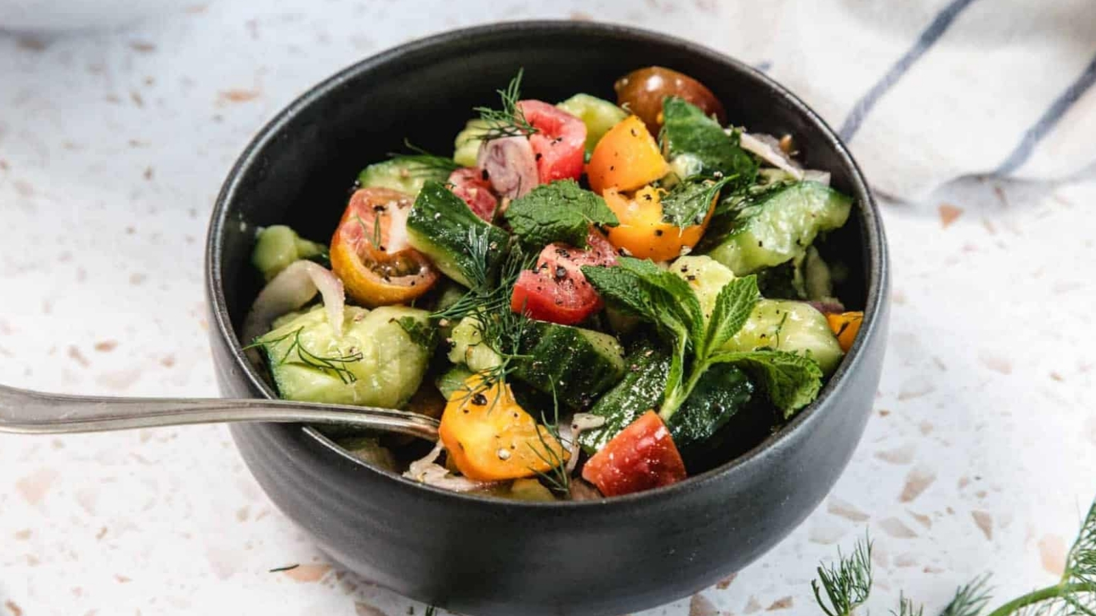 A black bowl filled with a salad made of sliced cucumbers, cherry tomatoes in various colors, fresh herbs, and a fork resting on the side. A white cloth with blue stripes is partially visible in the background.